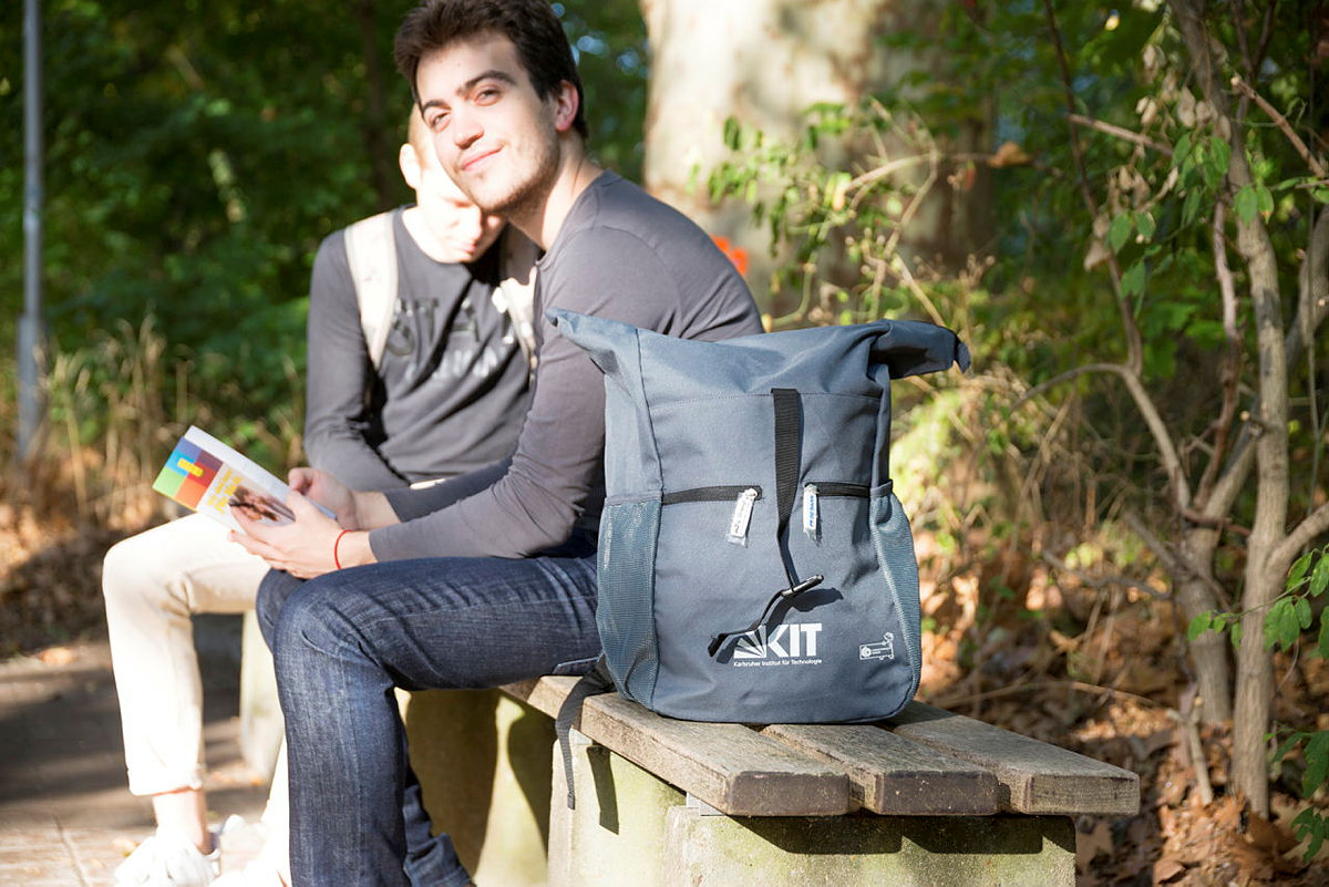 Young person sitting on a bench on Campus South, a KIT backpack beside
