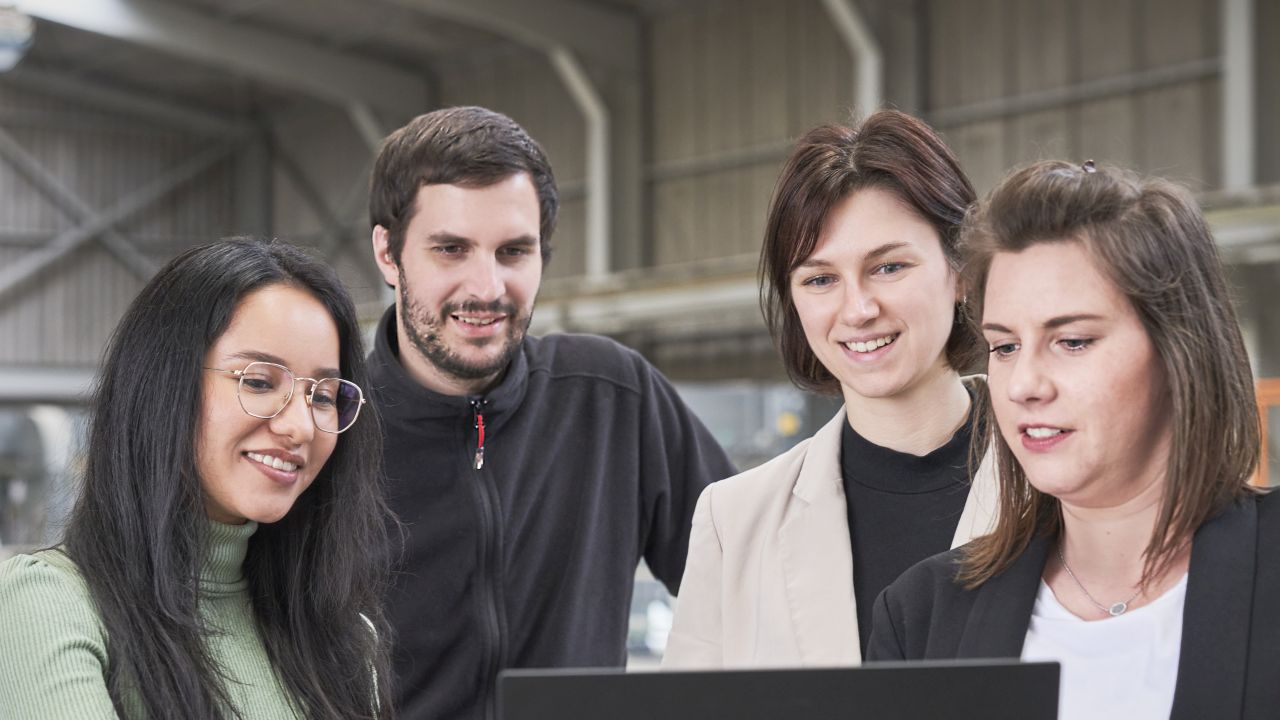Gruppe von vier jungen Berufstätigen schaut interessiert auf einen Laptop in einer technischen Umgebung.