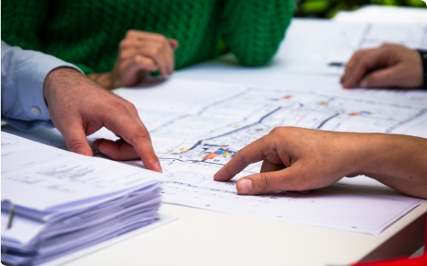 Mehrere Hände zeigen auf einen Bauplan auf einem Tisch.