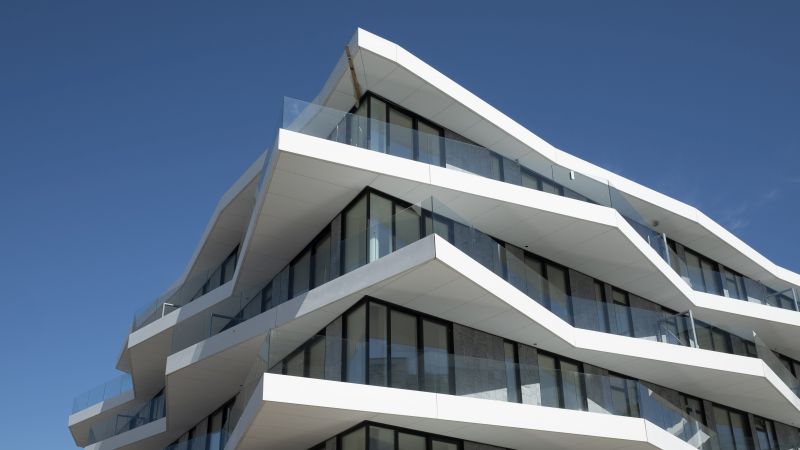 Moderner weißer Gebäudekomplex mit markanten, zickzackförmigen Balkonen vor blauem Himmel.