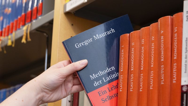 Hand greift in einem Bücherregal nach dem Buch „Methoden der Latinistik“ von Gregor Maurach.