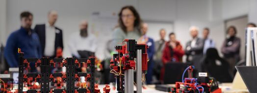 Im Vordergrund befinden sich technische Bauteile, die verkabelt sind. Im Hintergrund ist eine Menschengruppe unscharf zu erkennen. Eine Frau erklärt den Umstehenden, die Bauteile.