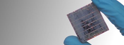 Eine blaue behandschuhte Hand hält eine kleine Solarzelle zwischen Daumen und Zeigefinger