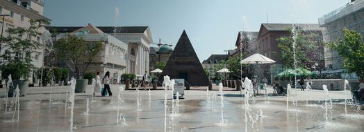Der Karlsruher Marktplatz im Sommer: Vor der Pyramide spritzen Wasserfontänen aus dem Boden, rechts und links stehen große Topfpflanzen auf dem hellen Pflaster.